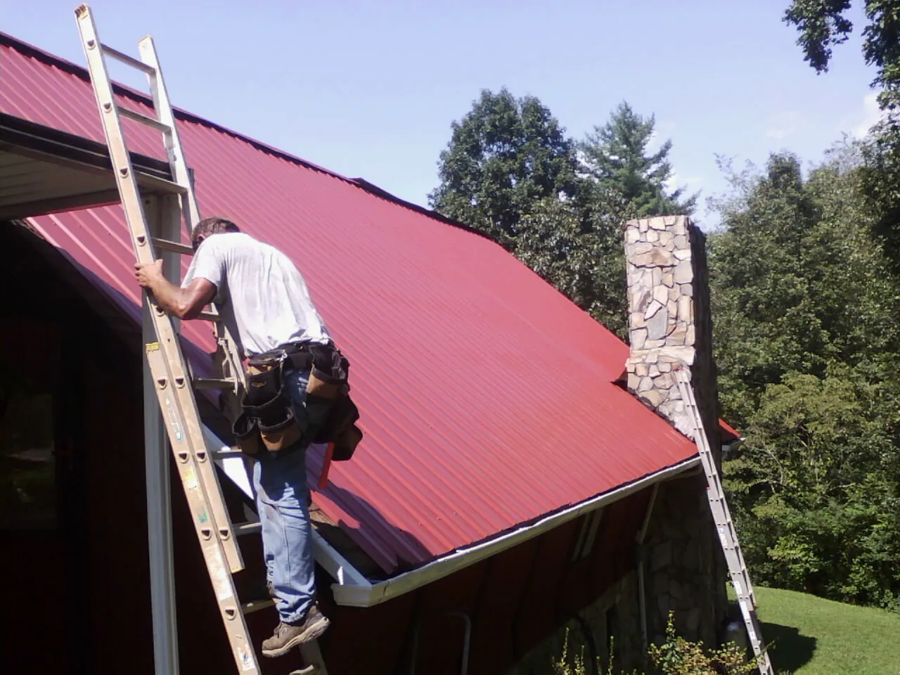 Roofer on a ladder beside a red metal roof and stone chimney — Soffit & Fascia Repair by Roofing Brothers in Wilson